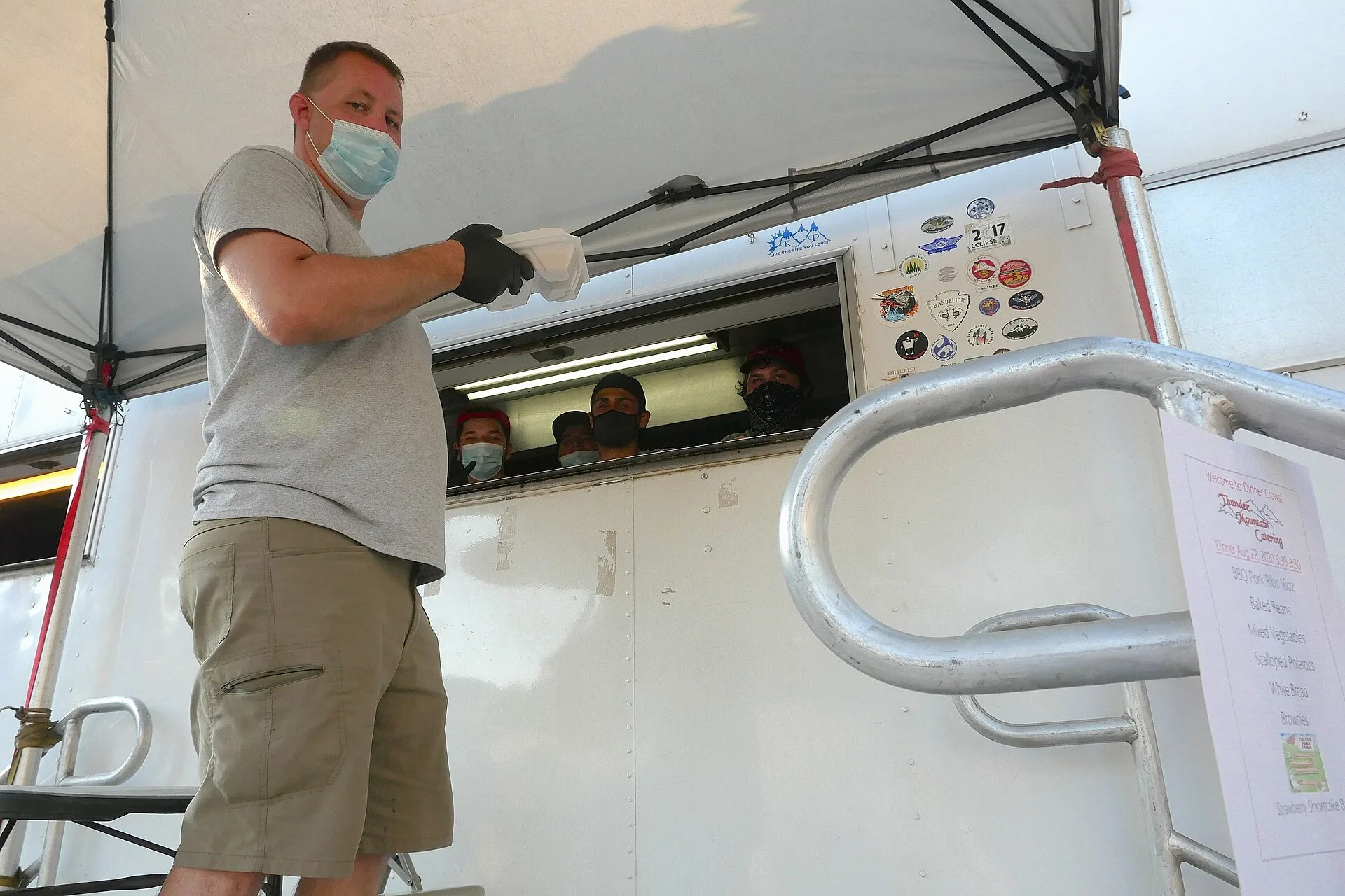 Temperature checks and face coverings are part of daily life in fire camp. Food service workers wear face coverings. Photo depicts three individuals peeking from a window in a food catering truck wearing face coverings. And one individual in a light blue face covering and gloves holding a togo conta. Food truck pricing: a block test for pitches and events